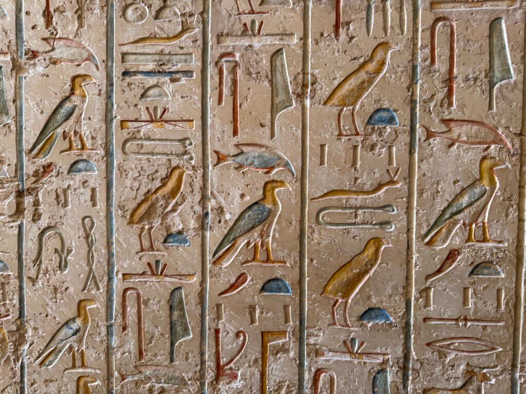 Detaljert utsnitt av egyptiske hieroglyfer skåret i en vertikal steinplate. Symbolene inkluderer blant annet en fugl, en sittende person og ulike geometriske figurer. Overflaten er slitt og ujevn, med spor av alder og erosjon.