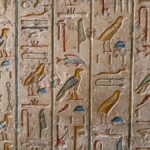Detaljert utsnitt av egyptiske hieroglyfer skåret i en vertikal steinplate. Symbolene inkluderer blant annet en fugl, en sittende person og ulike geometriske figurer. Overflaten er slitt og ujevn, med spor av alder og erosjon.