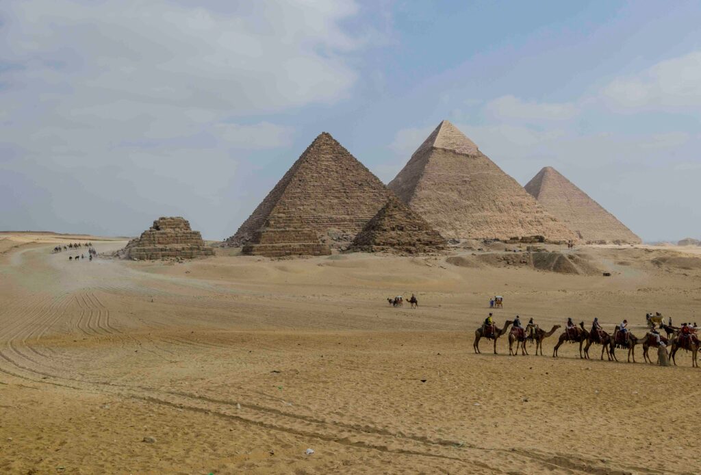 Foto av pyramidene på Giza-platået i Egypt, med flere store pyramider i bakgrunnen og en gruppe kamelryttere som krysser ørkensanden i forgrunnen.
