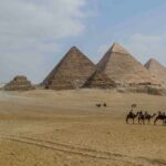Foto av pyramidene på Giza-platået i Egypt, med flere store pyramider i bakgrunnen og en gruppe kamelryttere som krysser ørkensanden i forgrunnen.