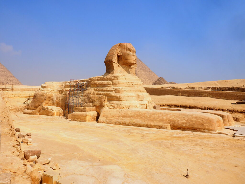 Foto av den store sfinxen ved Giza, en monumental steinskulptur med løvekropp og menneskehode, omgitt av ørkensand og med pyramider i bakgrunnen under en klar blå himmel
