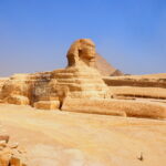 Foto av den store sfinxen ved Giza, en monumental steinskulptur med løvekropp og menneskehode, omgitt av ørkensand og med pyramider i bakgrunnen under en klar blå himmel