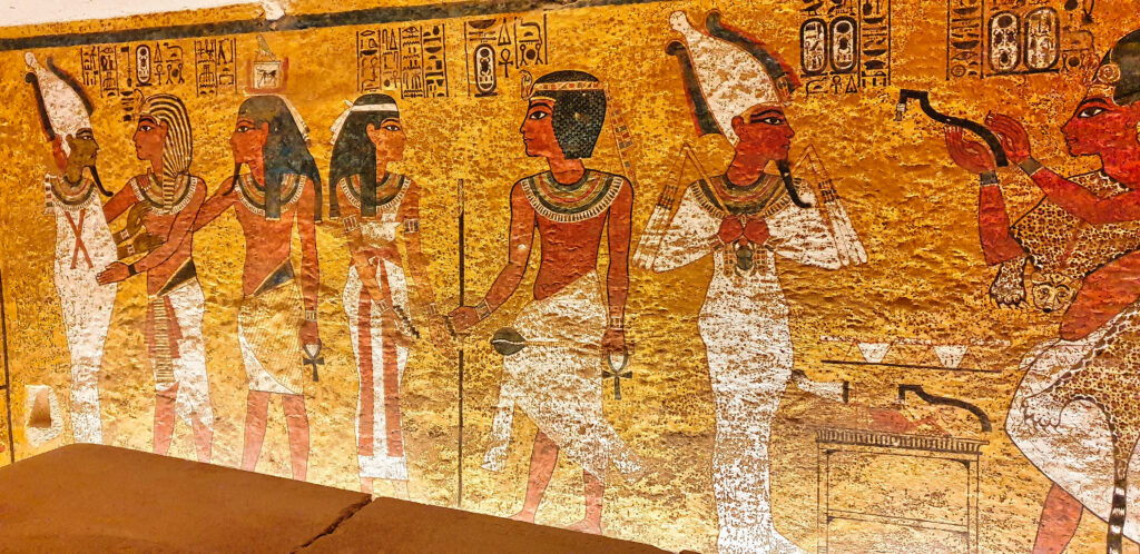 Fargerik veggmaleri i gammel egyptisk stil som viser flere stående figurer i tradisjonelle klær og hodeplagg, malt på en gyllen bakgrunn med hieroglyfer over.