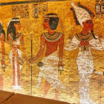Fargerik veggmaleri i gammel egyptisk stil som viser flere stående figurer i tradisjonelle klær og hodeplagg, malt på en gyllen bakgrunn med hieroglyfer over.