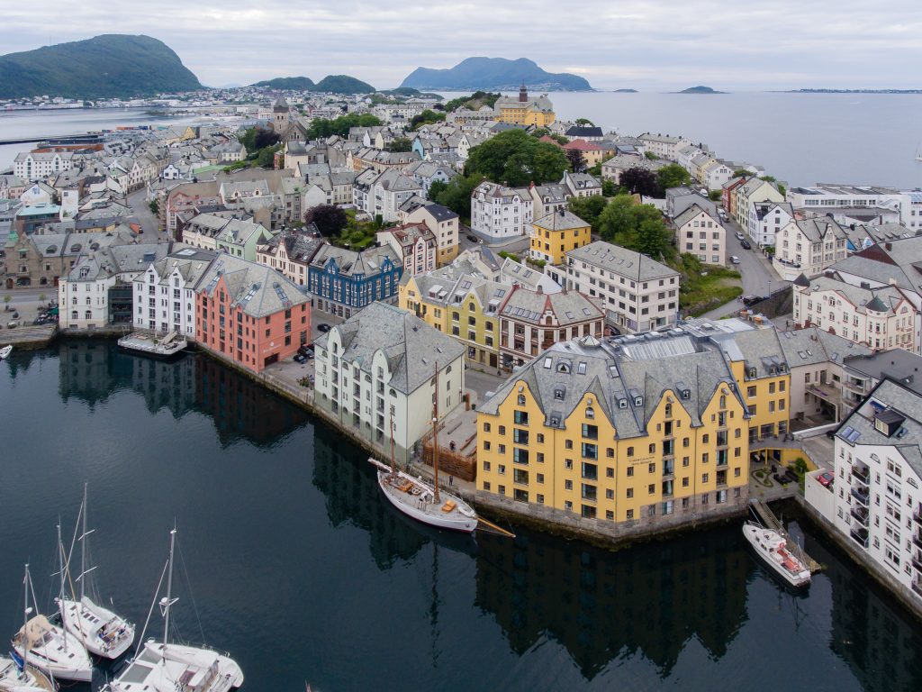 Fotografi av en by med murbygninger i flere etasjer. Bygningene er malt i gult, grønt, blått og rosa. Fasadene har buede linjer. Byen er omgitt av hav og fjell. 