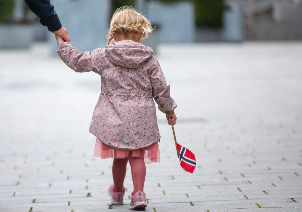 bilde av en liten jente som har på seg kjole, jakke og sko. hun bærer det norske flagget i hånda