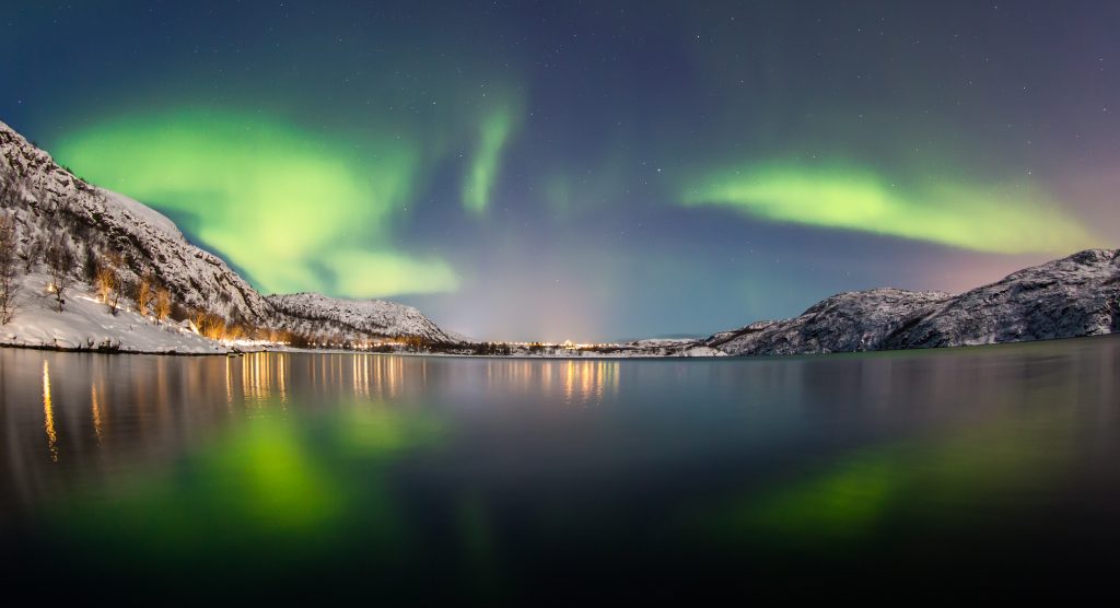 Fotografi av landskap med mye vann. Grønt og rosa lys slynger seg på himmelen og lyset speiler seg i vannet.