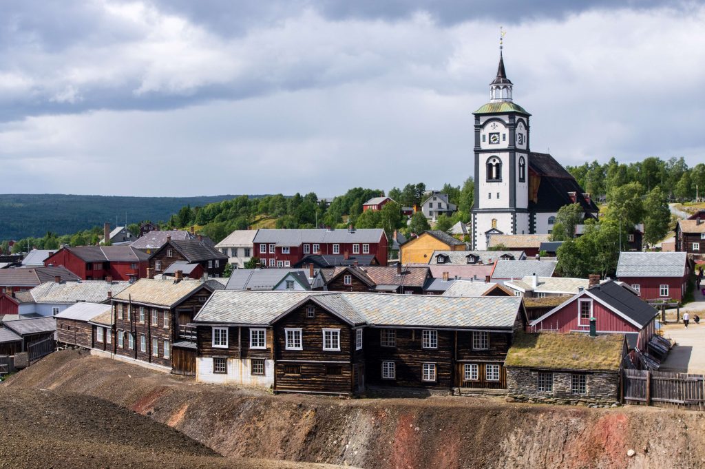 Fotografi av en liten by med en hvit trekirke og lave tømmerhus. I forgrunnen en store hauger med gruveslagg.