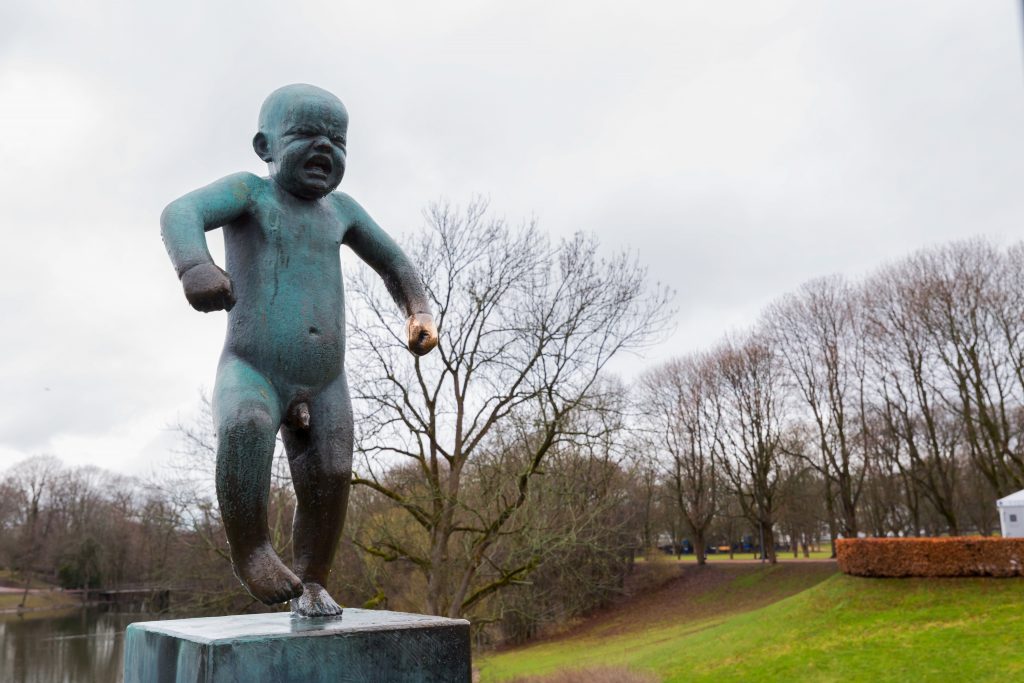 Fotografi av en bronsestatue som viser en liten gutt i midten av et raserianfall, med kroppen spent og ansiktet vridd i sinne. Bak statuen er det nakne busker,