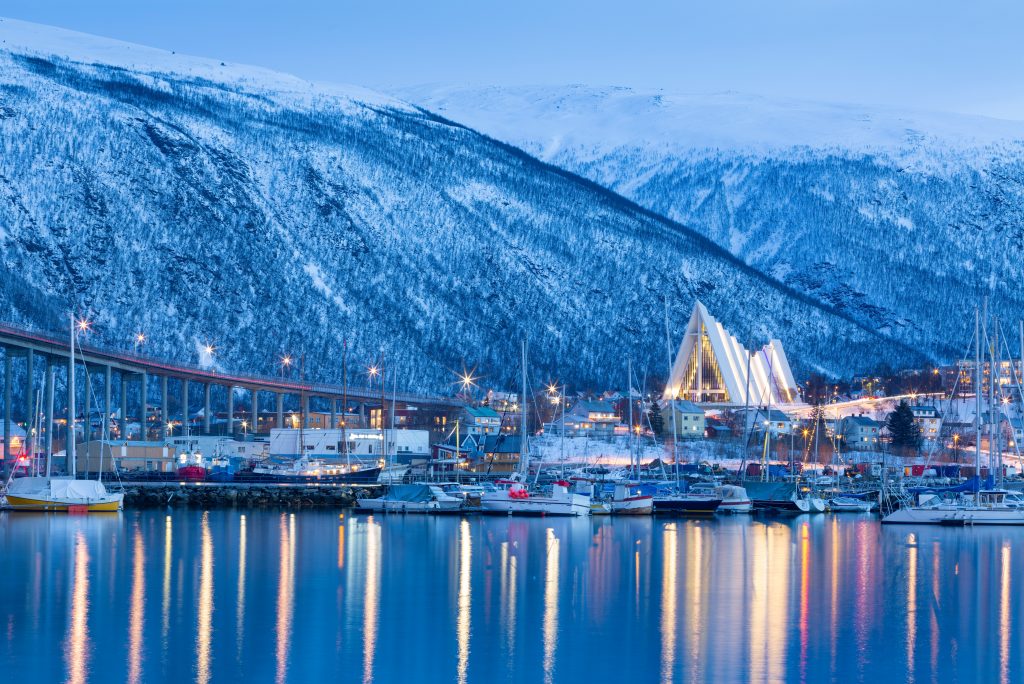 Fotografi av en by. Bildet domineres av en stor, hvit bygning som består av 11 trekantige deler i ulike høyder som er plassert inntil hverandre. Formen på bygget kan minne om isbreer og fjell. Det er vinter og snø. Lyset er blått.