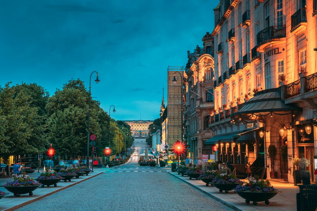 Kveldsbilde av en brosteinsbelagt gate med høye bygninger på høyre side og store trær på venstre side. I bakgrunnen er en gul murbygning med en opplyst altan med seks søyler.