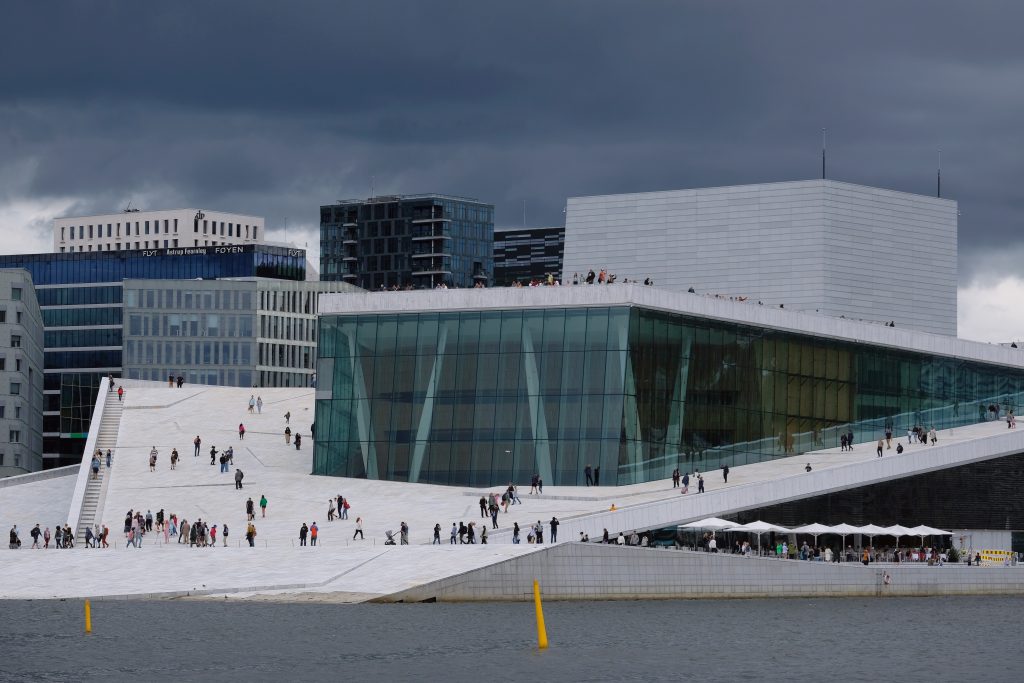 Et bilde av et markant bygg i hvit marmor og glass. Bygningen ligger ved vannkanten. Taket heller ned mot bakken, noe som gjør det mulig for folk å gå opp på det. På bildet er det mørkeblå skyer.