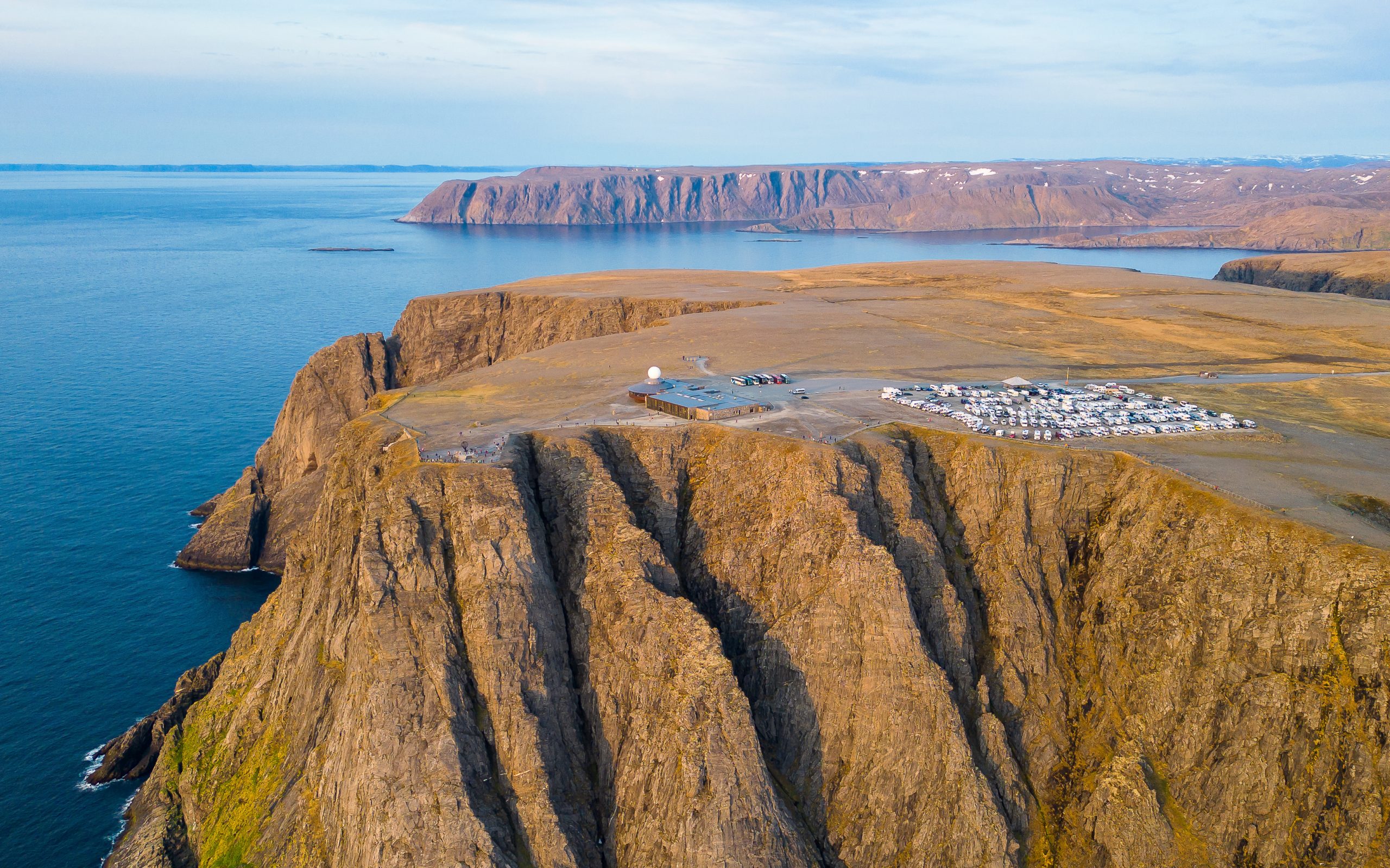 Oversiktsbilde av hav, nakent klippelandskap, noen bygninger og en parkeringsplass. Ved bygningene står et monument som er formet som en globe.