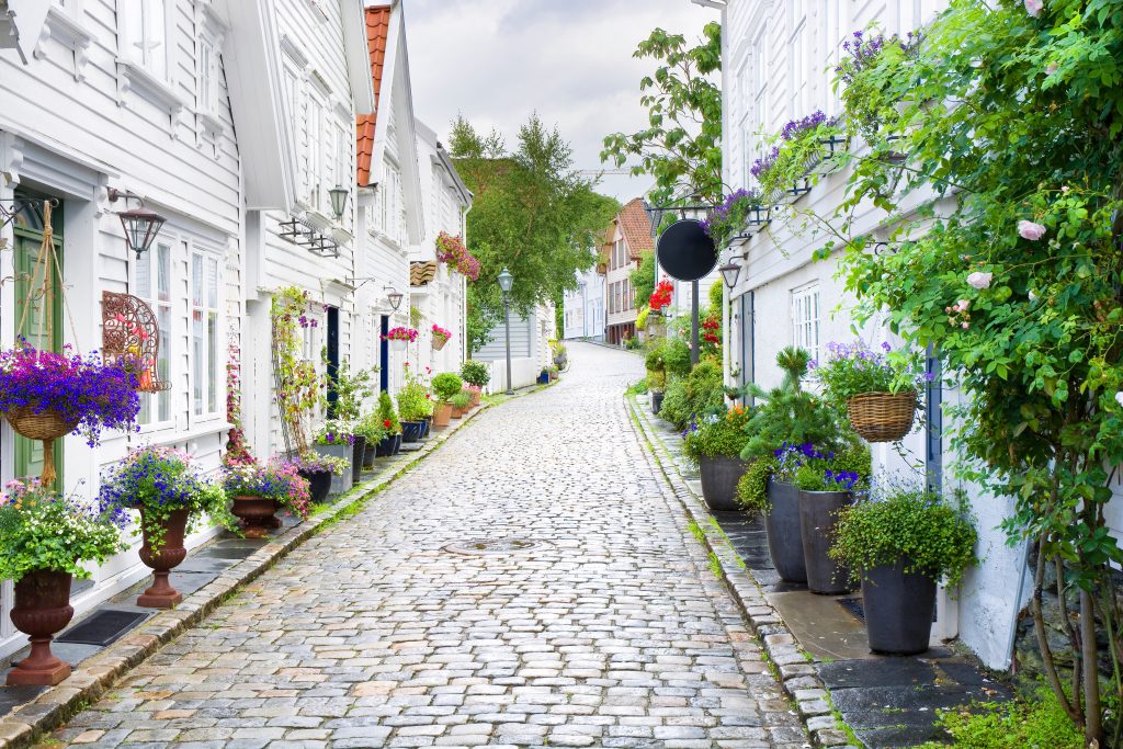 Fotografi av en smal, brosteinsbelagt gate. På begge sider av gaten er det hvite trehus og mange prydplanter.