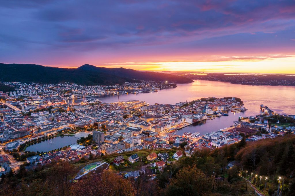 Oversiktsbilde av en by i solnedgang. vann og høye fjell i bakgrunnen.