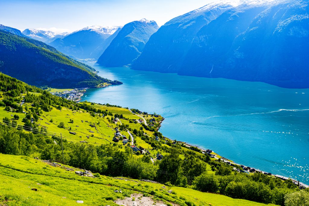 Fotografi av et landskap med vann, nakne fjell helt ned til vannet på den ene siden av vannet. På den andre siden er det grønne skråninger med gress og trær, veier og hus.