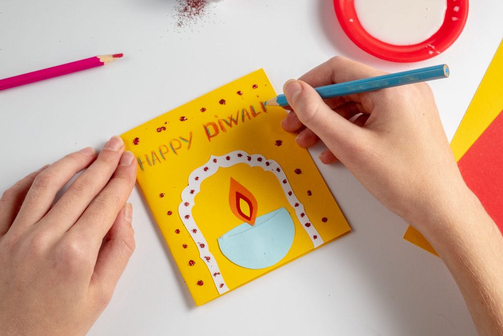 Happy diwali concept. A faceless child makes a postcard with a bowl of fire. Various materials for creativity - cardboard, glue, glitter, pencils