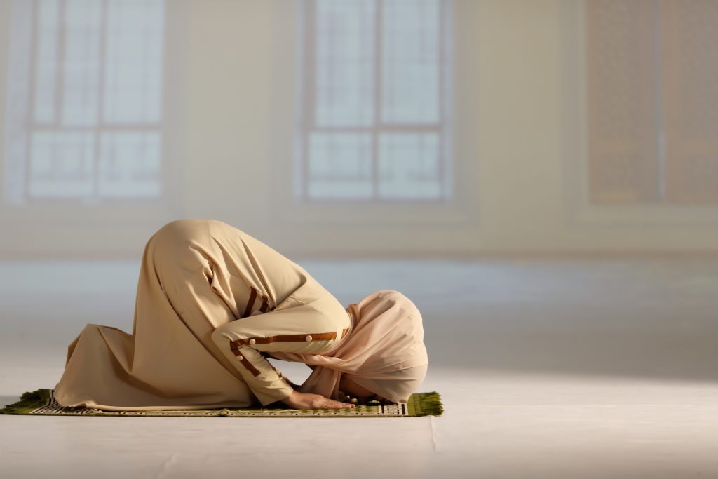 BIlde av en kvinne i lang beige kjole som bøyer seg i bønn mot Mekka. 