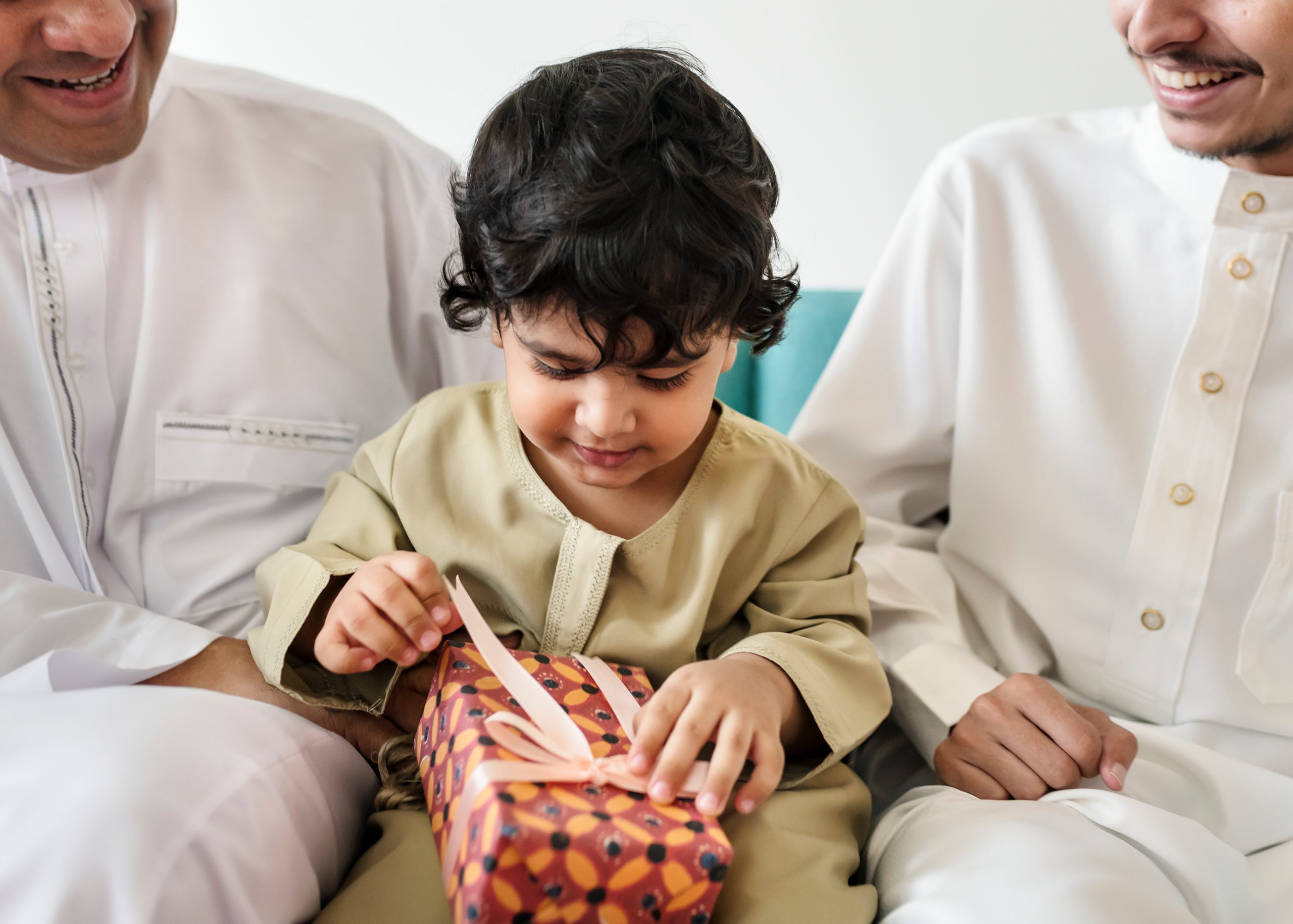 En liten gutt sitter i mellom to menn på en sofa og åpner en gave. Gaven er pakket inn i med beige sinkesløyfe og orange papir.