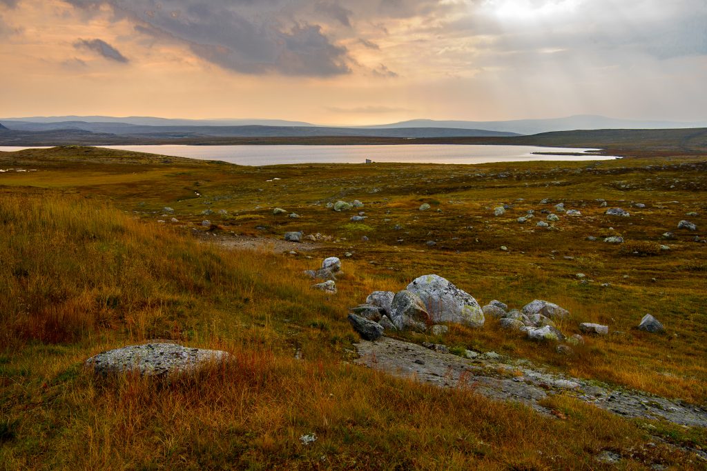 Fotografi av et naturområde som er ganske flatt med noen store steiner som stikker opp av brungrønn lyng. Det er en stor innsjø på bildet og hangt i det fjerne er det fjell i forskjellige blåfarger.