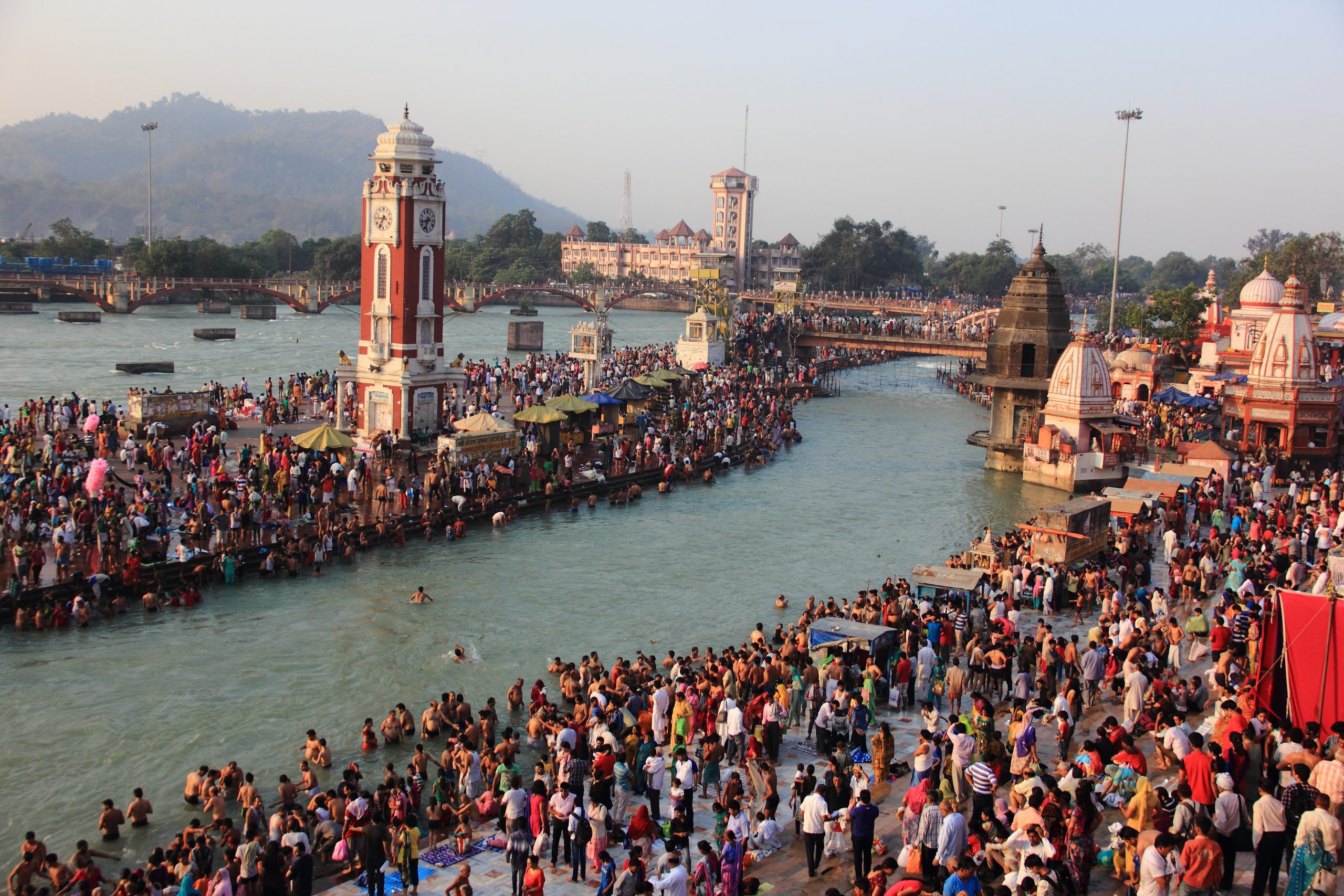 Mnge mennesker står langs kanten av Ganges. Noen bader og det er hinduistiske byggninger langs elvekanten