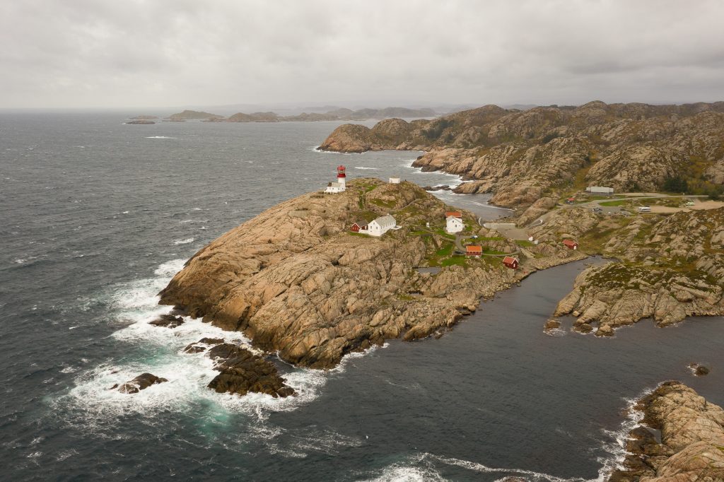 Oversiktsbilde av Lindesnes fyr. Bildet viser Lindesnes fyr, en hvit og rød bygning, plassert på en klippe ved kysten. Havet strekker seg ut i horisonten rundt fyret, og himmelen over er skyet.