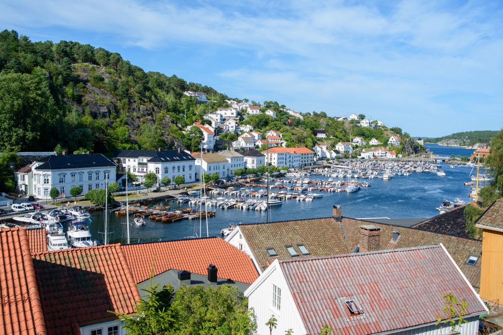 Fotografi av en by med hvitmalte trehus som ligger tett inntil hverandre langs vannkanten og oppover skråningen. På vannet ligger mange fritidsbåter , med grønne trær som bryter opp det hvite. Over byen er himmelen blå med noen spredte skyer.