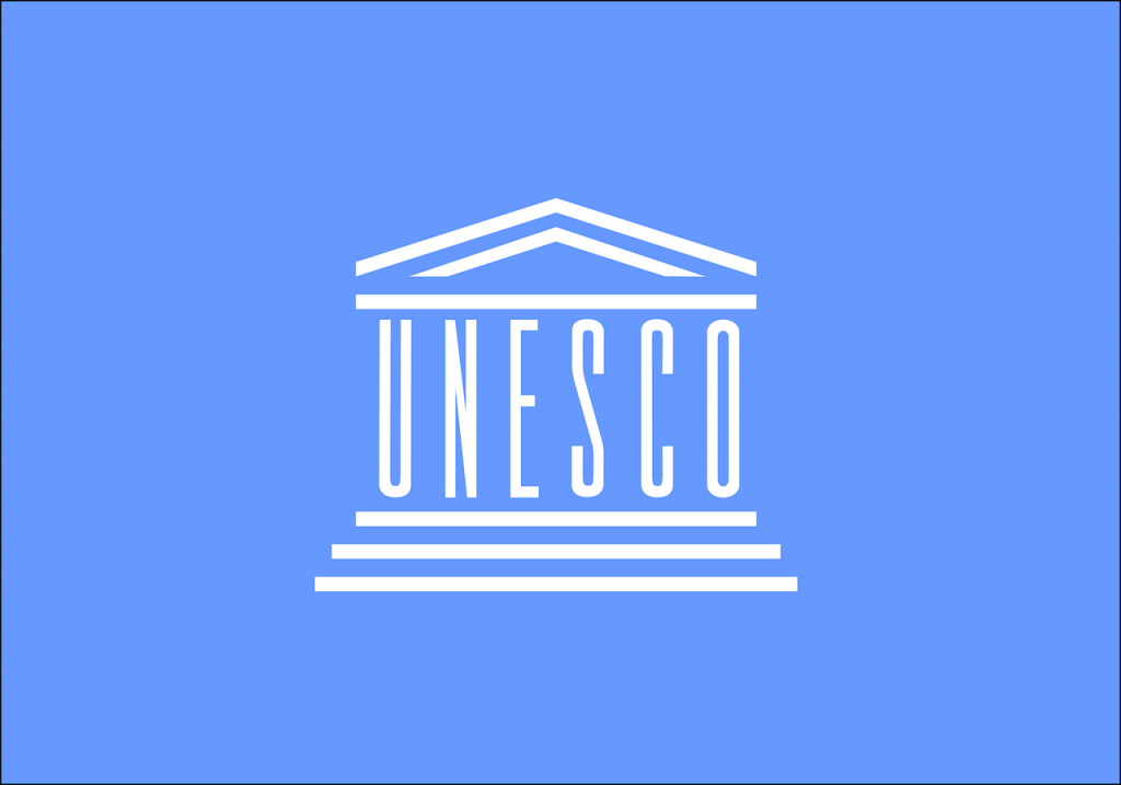 hvit tegning på lyseblå bakgrunn. Tegningen viser et bygg der bokstavebe UNESCO symoliserer søyler