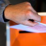 Fotografi av en hånd som holder en stemmeseddel delvis inne i sen orange stemmeurne
