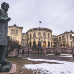 Fotografi av Stortinget, Norges nasjonalforsamling, sett fra utsiden. I forgrunnen står en statue av Johan Sverdrup.. Statuen viser Sverdrup i en stående posisjon, kledd i en lang frakk, med blikket vendt mot Stortinget.