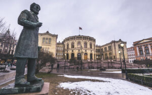 Fotografi av Stortinget, Norges nasjonalforsamling, sett fra utsiden. I forgrunnen står en statue av Johan Sverdrup.. Statuen viser Sverdrup i en stående posisjon, kledd i en lang frakk, med blikket vendt mot Stortinget.