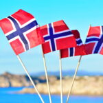 Fotografi av fire håndholdte norske flagg på bakgrunn som inneholder vann og berg.
